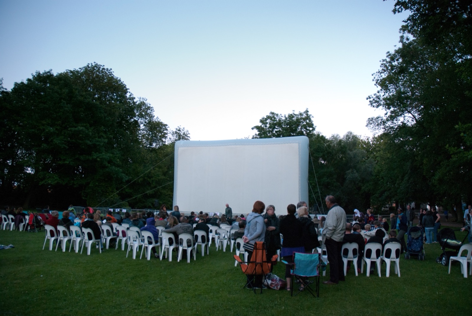 A giant inflatable cinema screen by Hallucine set up in a park, with the audience taking their seats at dusk